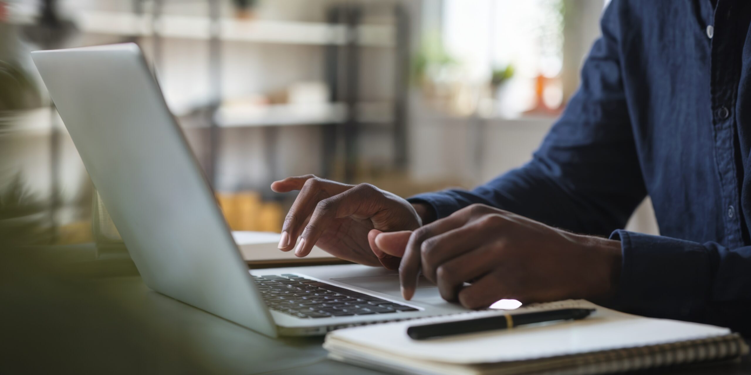 A man is typing on a laptop in front of a notebook and a cell phone. The laptop is open and the man’s hand is on the keyboard. The scene suggests that the man is working or studying