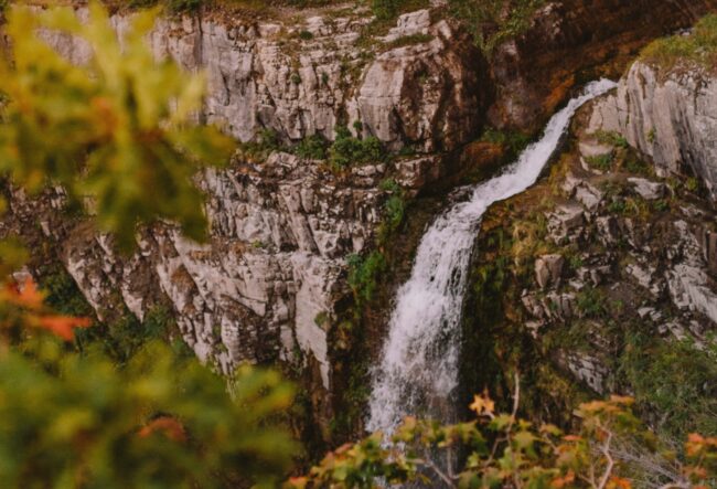 A scenic waterfall cascading down a rugged cliffside surrounded by autumn foliage, with colorful leaves framing the view of Stewart Falls in Utah.