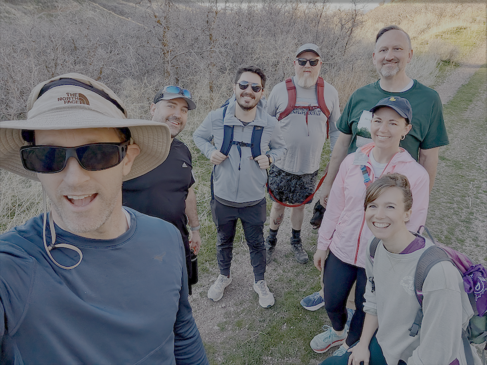 A group of 7 hikers standing on a trail surrounded by trees in springtime with blue skies overhead.
